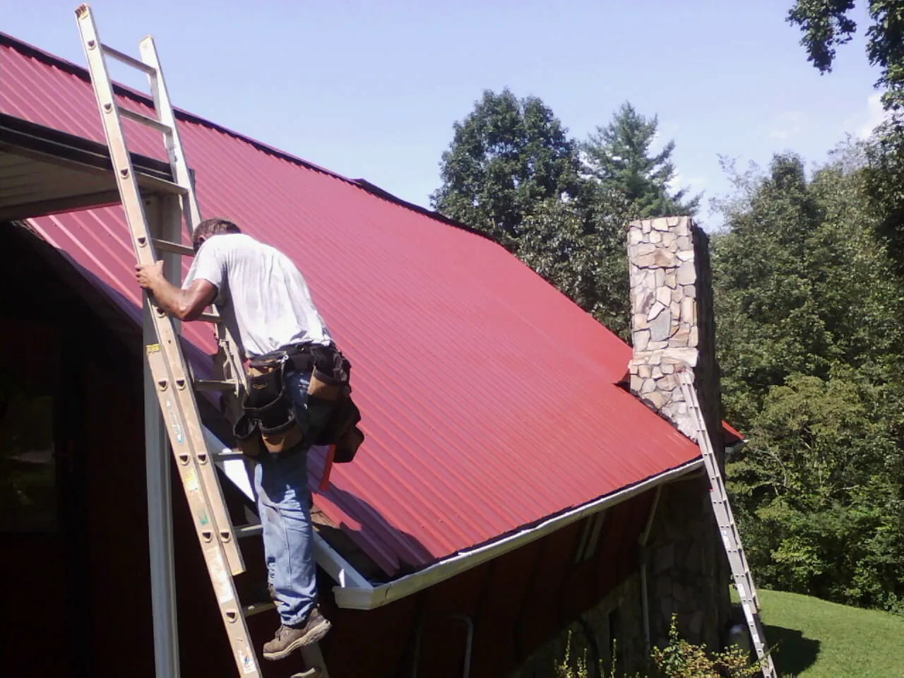 Roofer on a ladder beside a red metal roof and stone chimney — Soffit & Fascia Repair by Roofing Brothers in Mooresboro